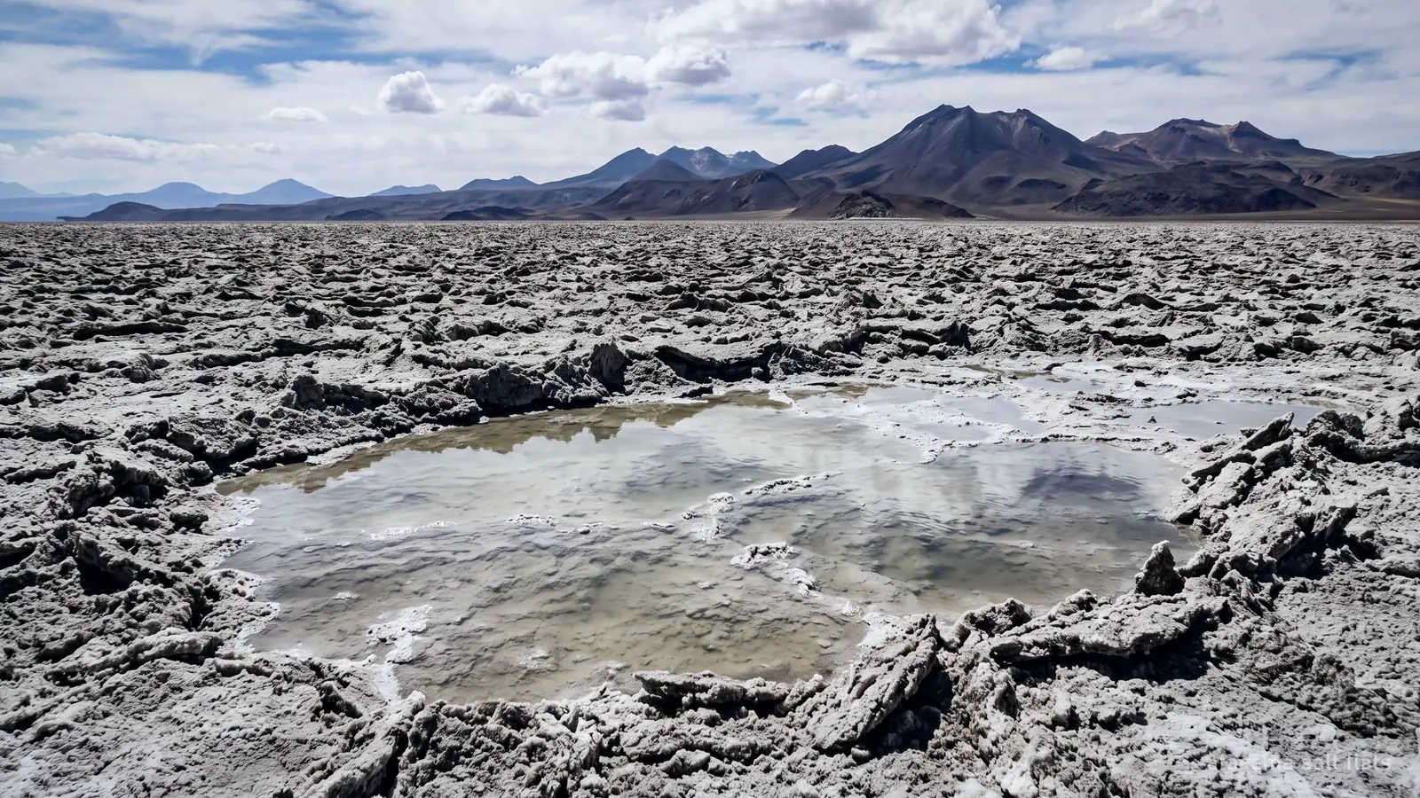 A rough grey salt crust stretching to distant volcanoes, with a shallow brine pool in the foreground