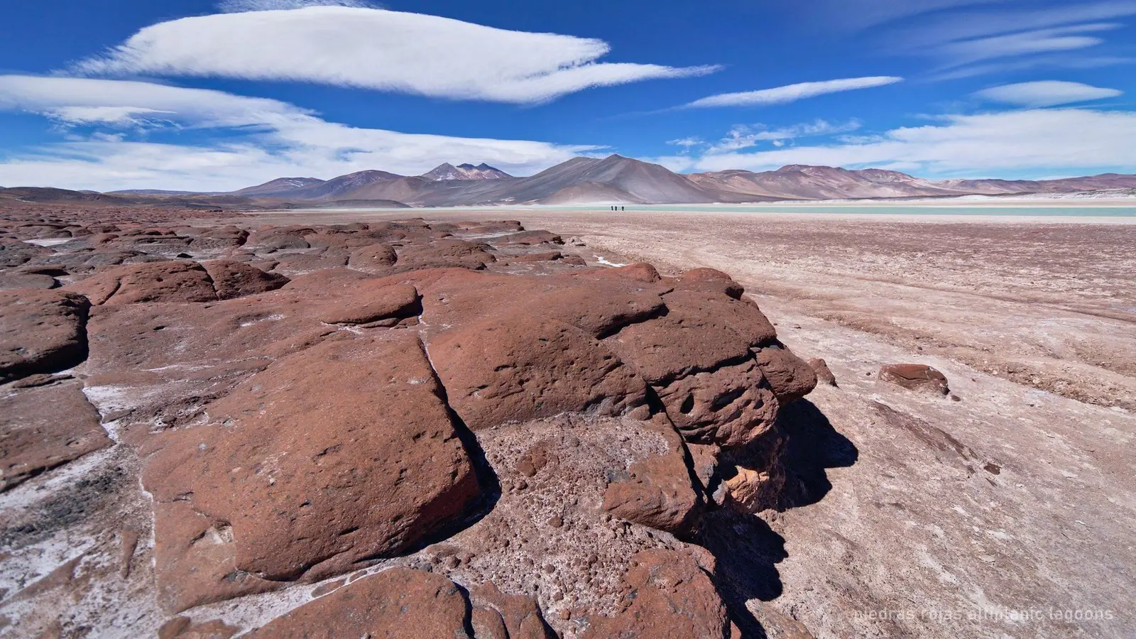 A high-altitude lagoon with red mineral rocks along the shore and a volcano behind